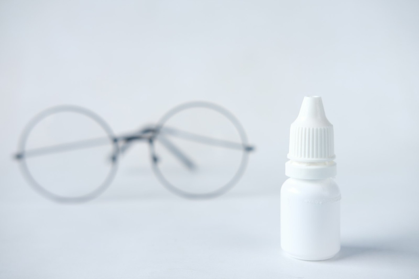 An eye-drop bottle beside a pair of glasses on a white table